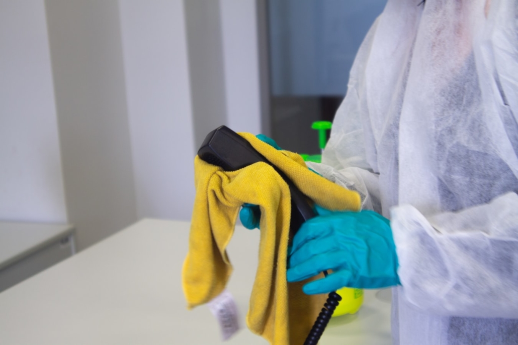 A cleaning person wearing blue gloves wiping down a phone in the office with a cloth