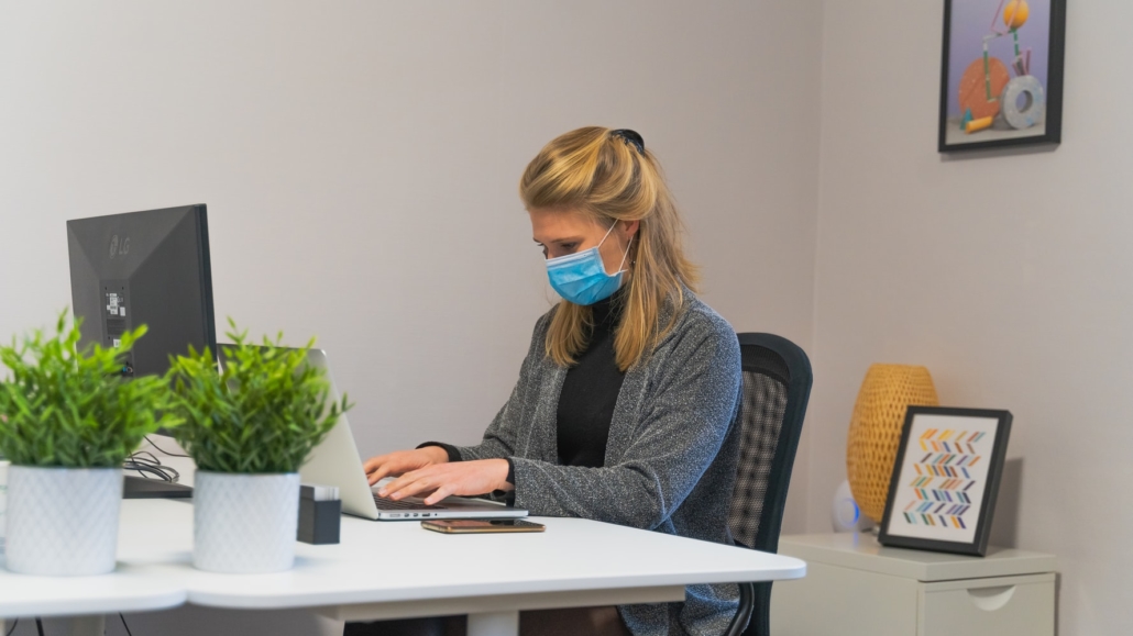 Blonde woman in the office wearing a mask at her desk adhering to social distancing guidelines